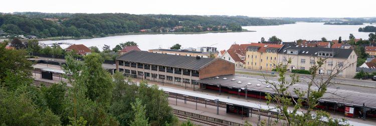 Oversigtsbillede af den tidligere posthusbygning med togskinnerne i forgrunden og Skanderborg Sø i baggrunden. 